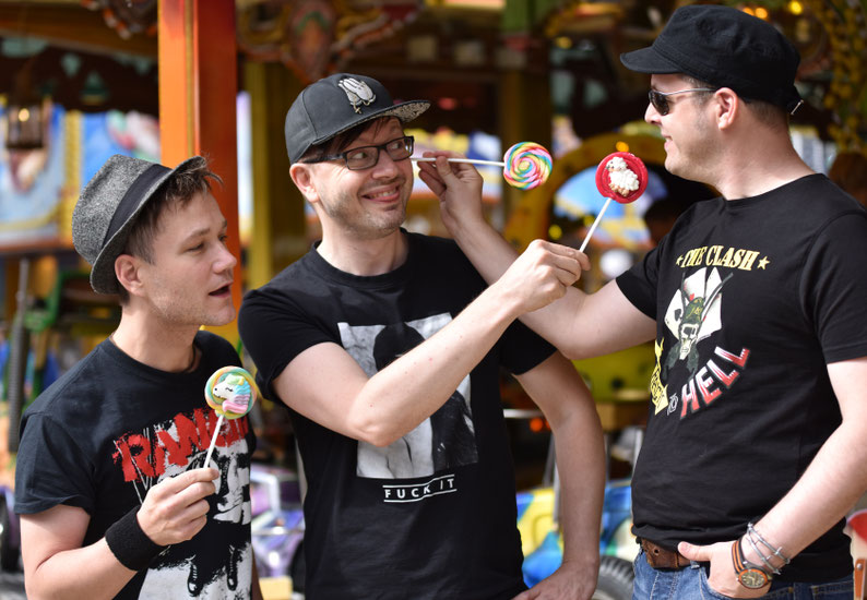 Anti Smile – drei Bandmitglieder mit bunten Lollis auf einem Jahrmarkt